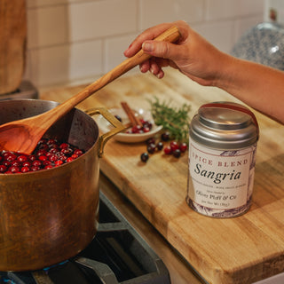 Person stirring a pot of berries with a wooden spoon, next to a can of Sangria spice blend on a kitchen counter.