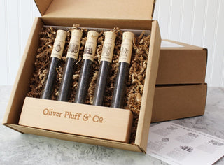 5 skinny clear tubes with corks holding loose leaf teas standing horizontally in brown wooden rectangular stand inside a brown box with brown stuffing paper