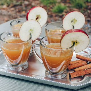 silver tray holding 4 clear glasses filled with tea, apple slices and cinnamon sticks. on a table outside