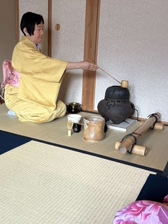 Japanese woman in yellow robe sitting on ground making tea with pots, cups and bamboo beside her