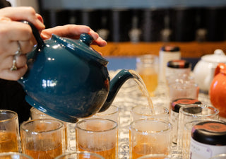 a teal teapot pouring tea into several clear glasses lined up on a tabletop