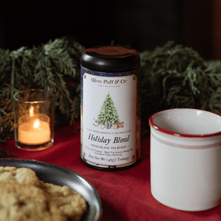 black tea tin with Christmas tree logo deside white mug, plate of cookies, candle and green garland on red table