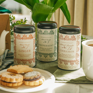 three black tea tins with different labels on table beside plate of shortbread cookies, green plant and white tea mug