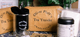 brown zipper pounch that reads 'oliver pluff's tea traveler charleston' next to clear glass with velvet bag inside of it