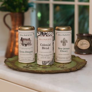 three gold tea tins in a row sitting on green plate on kitchen counter with coffee pot and pitcher blurred behind