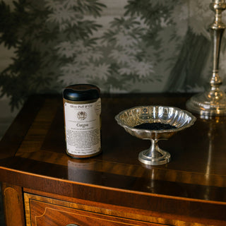 tin of tea with black lid on ornate wood tabletop next to silver dish filled with loose tea