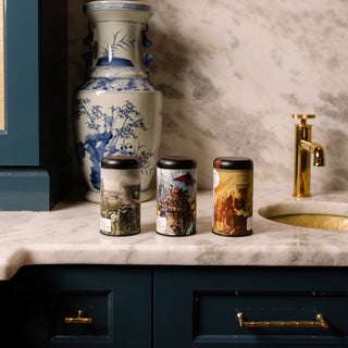 Three cylindrical tea tins with artistic designs on a marble countertop next to a decorative vase and gold faucet.