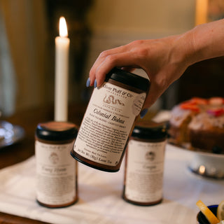 Hand holding a tin labeled 'Colonial Bohea' on a table with other cups and a cake.
