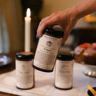 Hand holding a tea tin labeled 'Congou Loose' on a table with other cups and a candle.