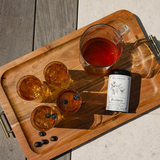Wooden tray with glasses of amber liquid, a jar, and a bottle labeled 'Blueberry' on a wooden surface.