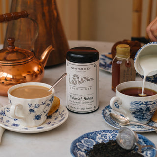 a black tea tin with white label on white tablecloth, surrounded by filled blue and white teacups