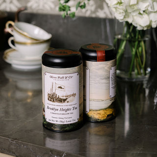 Two tea canisters with vintage designs on labels, placed on a reflective surface with a blurred background.