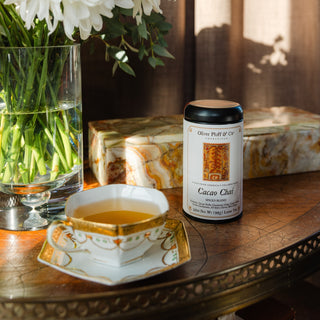 tea tin with white label that reads cacao chai loose, sitting on wooden tabletop next to filled teacup and marble box