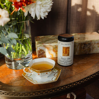 cacao chai teabags tea tin with white label and black lid on wooden tabletop, surrounded by floral arrangement and a teacup filled with brewed tea
