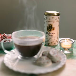 christmas blend coffee tin on tabletop surrounded by cup of hot, steaming coffee, cookies and peppermints