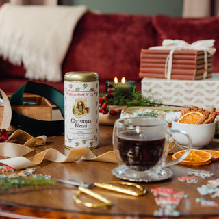 a tin of christmas coffee sitting on a wooden tabletop surrounded by gift wrapping materials and a cup of brewed coffee