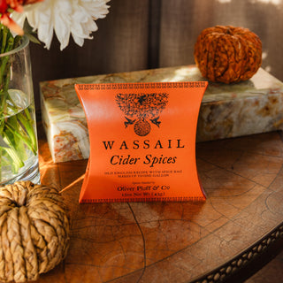 orange pillow box that reads 'wassail cider spices' on wooden tabletop with woven pumpkins in the foreground and background