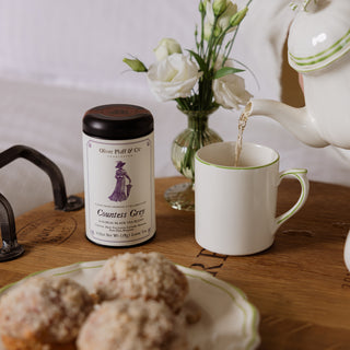 Tea being poured into a cup with a jar of tea and muffins on a wooden table.