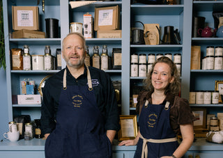 Two people wearing aprons standing in front of a shelf with various items.