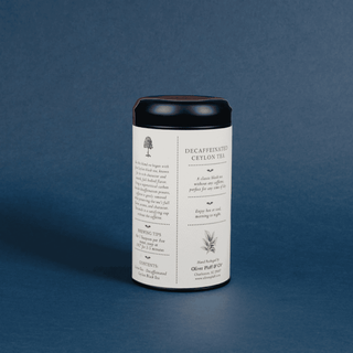 A cylindrical metal canister of Olive Orchard Co. Decaffeinated Ceylon Tea, featuring a white label with black text and illustrations, topped with a black lid, set against a textured dark blue background.