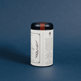 A white cylindrical tea tin with a black lid and a brown leather loop, standing on a dark blue background. The label features elegant black typography and botanical illustrations.