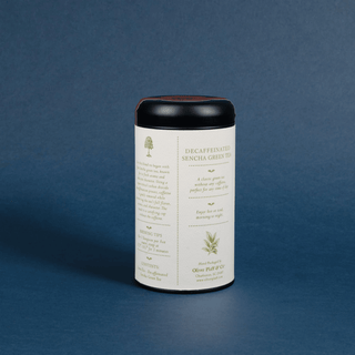 A white cylindrical tin of Decaffeinated Sencha Green Tea with a black lid, set against a textured dark blue background.