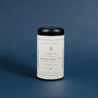 A cylindrical canister of Oliver Pluff & Co. Decaffeinated Sencha Green Tea, featuring an off-white label with green botanical illustrations and a black lid, set against a solid dark blue background.