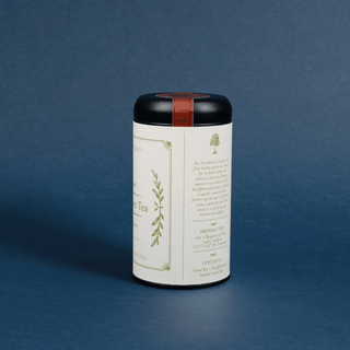 A cylindrical metal tea tin with a white label and a black lid, featuring a thin copper-brown band around the lid, set against a solid dark blue background.