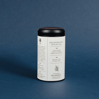 A cylindrical metal tin of Olive Puff & Co. Decaffeinated Ceylon Tea, featuring a white label with black text and illustrations, a black lid, and a dark brown tea fill visible at the top, set against a textured blue background.