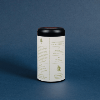 A cylindrical canister of decaffeinated sencha green tea, featuring a cream-colored label with green text and illustrations, a black lid, and a dark red inner rim, set against a textured dark blue background.