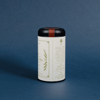 A cylindrical metal tea tin with a black lid and a warm brown leather band, standing upright on a dark blue textured background. The label is off-white with elegant gold botanical illustrations and text.