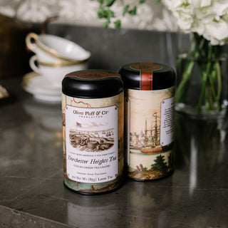 Two tea canisters with decorative labels on a dark surface, with a blurred background of a tea set and flowers.