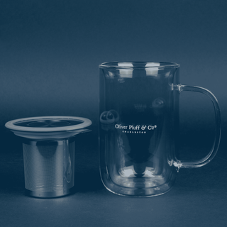 clear glass mug that reads 'oliver pluff & co' with stainless steel brewing basket sitting next to it with grey lid on blue background