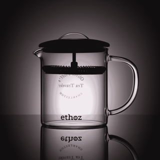 Clear glass teapot with black lid and handle on a reflective surface