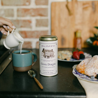 tin of green dragon coffee with white label and gold lid on black countertop. milk is being poured into a teal blue coffee cup behind it, with pastries on a plate in the foreground