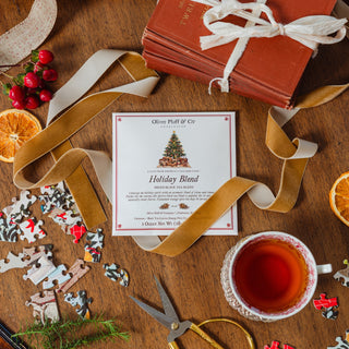 A flat-lay of a 'Holiday Blend' scented candle card by 'Olivia Puff & Co' on a rustic wooden table, surrounded by festive props including a cup of tea, scissors, jigsaw puzzles, red berries, orange slices, and a wrapped book.