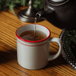 Tea being poured from a teapot into a white mug with a red rim on a wooden surface.