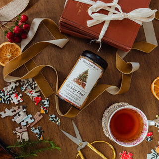 holiday blend tea tin laying flat on a wooden tabletop surrounded by gift wrapping materials, puzzle pieces, and a brewed cup of tea