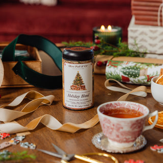 holiday blend tea tin on wooden table top with a cup of brewed tea in the foreground and wrapping materials surrounding it