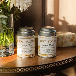 orange clove and lemon ginger hot toddy tins on wooden tabletop with marble box and floral arrangement in the background