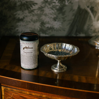 a tin of gunpowder loose tea on a wooden tabletop, next to a silver dish filled with loose tea