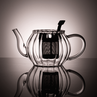 Clear glass teapot backlit to show interior infuser on a reflective surface.