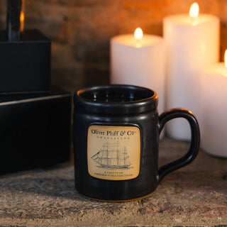 black barrel style mug that reads oliver pluff and co with ship design with lit candles in the background