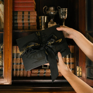 black gift set box tied with black ribbon in front of wooden bookshelf holding old books