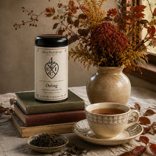 A tea tin labeled 'Oolong' stands on stacked books beside a cup of tea and dried flowers in a vase, set on a table by a window.
