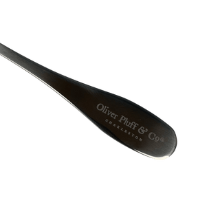 A close-up of a dark metal spoon head with engraved silver text reading 'Oliver Pluff & Co' and 'CHARLESTON' on a solid green background.