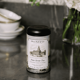 Tea canister labeled 'Oliver Pluff & Co' on a dark surface with floral and ceramic items in the background