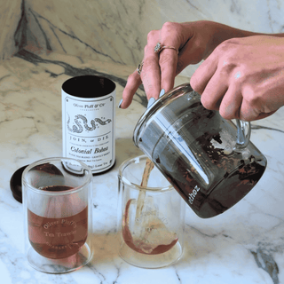 Person pouring liquid from a glass container into a jar on a marble surface with a can labeled 'Olive Leaf & Co' in the background.