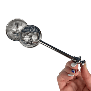 A hand holding a polished stainless steel double-sphere strainer with a long handle, set against a solid dark green background.
