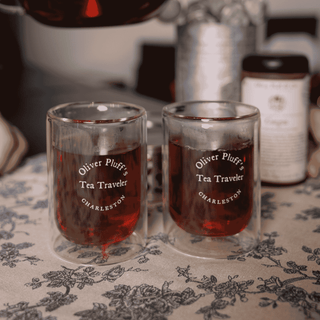 Two glass cups labeled 'Oliver Pluff's Tea Traveler' on a floral tablecloth.